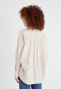 Camicia oversize a righe con strisce verticali beige e marroni, tessuto leggero, maniche con bottoni e vestibilità rilassata.