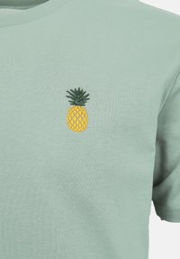 Φωτεινό πράσινο κοντομάνικο μπλουζάκι με μικρό κίτρινο κεντημένο ανανά και πράσινα φύλλα στην αριστερή πλευρά του στήθους.