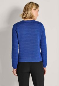 Vrouw met kort blond haar, gekleed in een gestructureerde blauwe lange mouwen trui en zwarte broek, van achteren afgebeeld tegen een effen muur.