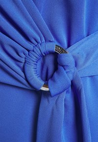 Modrý textilní oděv s uzlovým designem a stříbrným kruhovým kroužkem. Textura je hladká s mírným elastickým efektem. Detailní záběr.