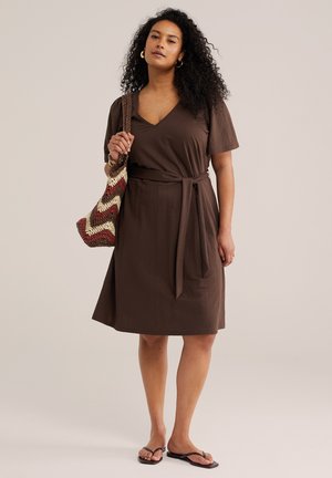 Femeie cu păr creț, purtând o rochie maro cu curea, ținând o geantă țesută cu model chevron și purtând papuci negri.