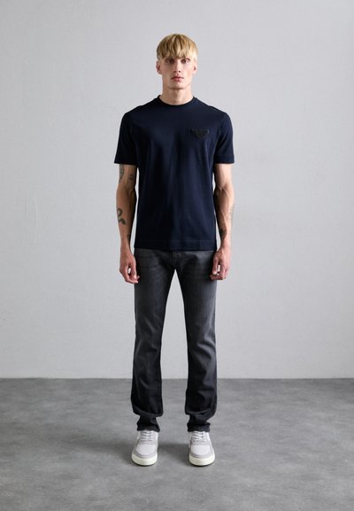 Navy cotton t-shirt with a crew neck and a small black logo on the chest, paired with dark gray jeans and gray sneakers with white soles.