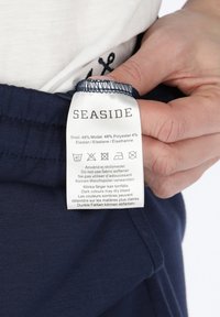 Navyblauwe stoffen label met onderhoudsinstructies en samenstelling: 48% modal, 48% polyester, 4% elastaan. Merkennaam: Seaside.