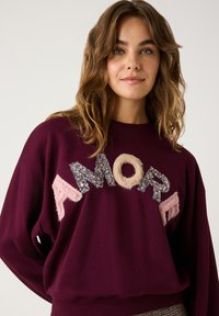 Junge Frau mit welligem Haar, die einen kastanienbraunen Pullover mit den Buchstaben "AMORE" aus Pailletten und flauschigen Texturen auf einem einfarbigen Hintergrund trägt.