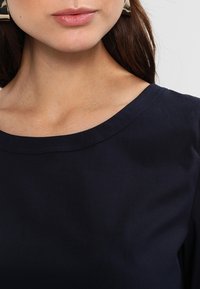 Mörkblå topp med rund halsringning, slät yta och korta ärmar. Noterbart detaljer inkluderar en lätt lyster på tyget.