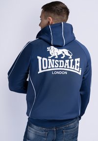 Mørkeblå hættetrøje med hvide detaljer, der har et stort løvegrafik og teksten "Lonsdale London" på ryggen, lavet af blødt stof.