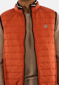 Gilet matelassé orange avec un col haut, doté d'une fermeture éclair noire et d'accents bleu marine, porté par-dessus un pull en tricot beige clair.