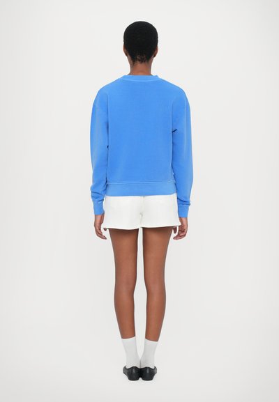 Μπλε βαμβακερό φούτερ με ριμπ μανσέτες και στρίφωμα, φορεμένο με λευκές βερμούδες. Το ύφασμα έχει απαλό υφή και άνετη εφαρμογή.