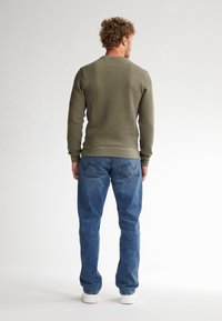 Pullover verde oliva con texture e collo rotondo, abbinato a jeans di denim blu chiaro e sneakers bianche. Design semplice e casual.