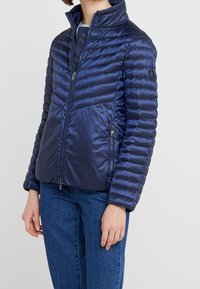 Navy puffer jacket with a fitted design, featuring horizontal quilted sections, zippered pockets, and a high collar. Smooth, shiny texture.