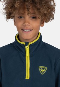 Pullover in pile blu con zip e colletto gialli. Presenta un patch con il logo Rossignol sul petto. Texture morbida e design con zip.