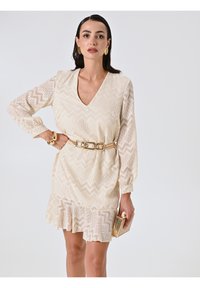 Robe à col en V et manches longues couleur crème avec une texture en zigzag. Dotée d'un ourlet à volants et d'une ceinture dorée. Complétée par une pochette dorée.