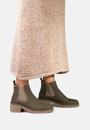 Bottes en daim vert olive arrivant à la cheville avec des empiècements latéraux élastiques beiges, portées avec une longue jupe beige tricotée.
