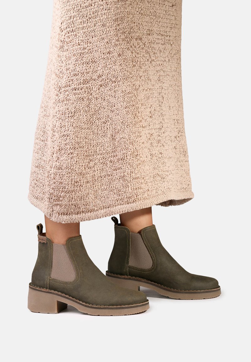Bottes en daim vert olive arrivant à la cheville avec des empiècements latéraux élastiques beiges, portées avec une longue jupe beige tricotée.