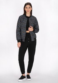 Zwarte bomberjack met een kleurrijk abstract patroon, geribbelde manchetten en zoom, gedragen over een zwart crop top en zwarte joggers, met zwarte sneakers.