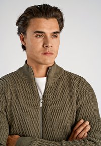 Olijfgroene gebreide cardigan met rits en een gestructureerd diamantpatroon en een geribde kraag, gedragen over een wit shirt, armen gekruist.
