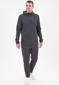 Felpa con cappuccio grigio scuro, con zip frontale a metà e tasca a marsupio, abbinata a pantaloni jogger coordinati e sneaker bianche. Tessuto morbido, design semplice.