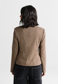 Sacou cu carouri maronii, cu o croială ajustată, având rever în formă de notă și mâneci lungi, asortat cu pantaloni din denim închis. Detalii de țesătură vizibile.