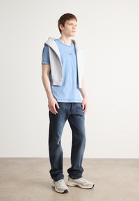 T-shirt azul claro com logotipo, hoodie cinza drapeado sobre os ombros, jeans escuros e ténis cinzentos com detalhes claros. Tecido suave, ajuste casual.