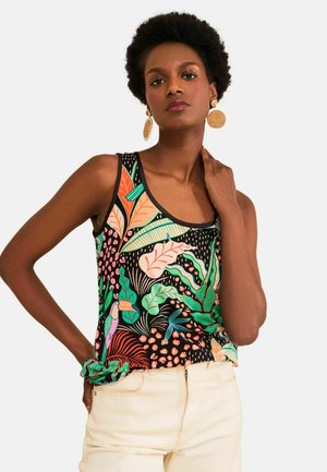 Woman with natural hair wearing large round woven earrings and a colorful tropical print sleeveless top with light-colored pants.
