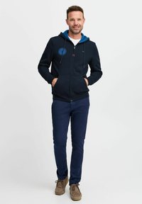 Felpa con zip blu marina, con interno cappuccio blu, tasche frontali e dettaglio del logo. Abbinata a pantaloni blu scuro e scarpe in suede marrone.