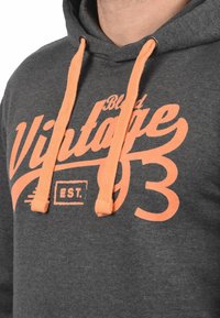Grijze hoodie met oranje koordjes en grote grafische tekst "Blend Vintage 93 EST." De stof lijkt zacht en heeft een casual ontwerp.