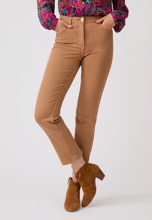 Donna con pantaloni slim fit color cuoio, stivaletti marroni in camoscio e una camicetta colorata a fiori con maniche lunghe, in piedi con le mani nelle tasche.
