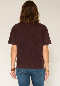 Femme aux cheveux bruns ondulés portant un t-shirt court noir délavé, un jean bleu et des bracelets dorés et marron, vue de dos.