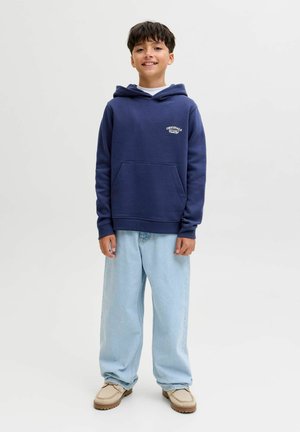Felpa con cappuccio blu navy con tasca frontale e logo, abbinata a jeans baggy azzurri e scarpe beige, in piedi davanti a uno sfondo neutro.