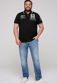 Black polo shirt with yellow accents, features printed design elements. Paired with light blue jeans and white sneakers. Relaxed fit.