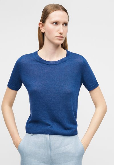 Άτομο με ίσια, ανοιχτό καστανά μαλλιά που φοράει μπλε πλεκτό μπλουζάκι με κοντά μανίκια και ανοιχτό γκρι παντελόνι, με τα χέρια στις τσέπες, κοιτάζοντας δεξιά.