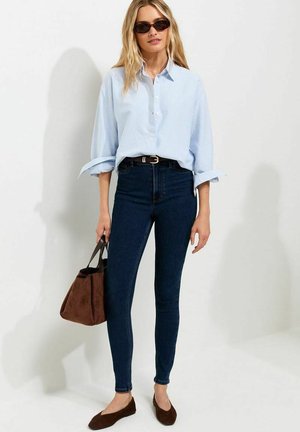 Vrouw draagt lichtblauw overhemd met knopen, donkere skinny jeans, bruine platte schoenen, zonnebril, houdt een bruine suède handtas vast tegen een witte achtergrond.