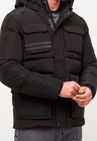 Schwarze Daunenjacke mit Reißverschluss und Knopfverschluss, großen Vordertaschen und einer Kapuze. Strukturierten Stoff mit horizontalen Streifen auf der Brust.