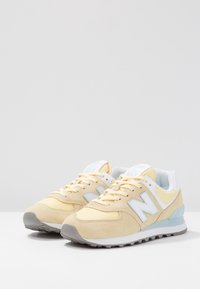 Gule sneakers i ruskind og mesh med lyseblå detaljer, med en grå gummisål og en fremtrædende hvid "N"-logo på siderne.