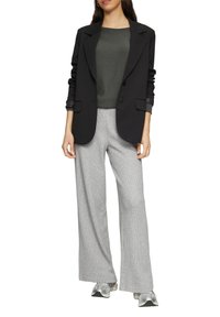 Schwarzer Blazer mit Revers und zwei Knöpfen, kombiniert mit einem dunkelgrauen Oberteil und hellgrauen karierten weiten Hosen. Sneaker runden das Outfit ab.
