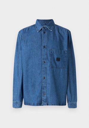 Camisa de mezclilla en azul oscuro con cuello abotonado, bolsillo en el frente y parche negro con logo. La tela es liviana con una textura suave.