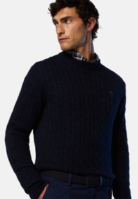 Maglione navy a trecce con scollatura rotonda e motivo testurizzato. Presenta polsini a costine e un piccolo logo. Indossato sopra una camicia a quadri.