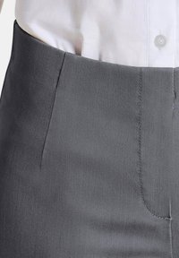 Grijze op maat gemaakte broek met een gladde textuur, details van voorkruiszakken en subtiele stiksels langs de tailleband, gecombineerd met een witte blouse.