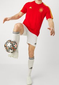 Rode Adidas voetbalshirt met gele strepen, witte shorts, witte sokken met zwarte strepen, en een zwarte voetbal met een sterpatroon.