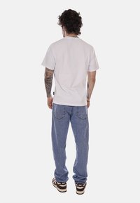 Uomo con capelli ricci e tatuaggi, visto di spalle, che indossa una t-shirt bianca semplice, jeans blu e scarpe Nike marroni.