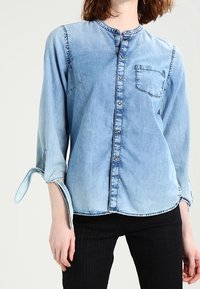 Camicia in denim azzurro chiaro con colletto alla coreana, chiusura frontale con bottoni, taschino sul petto e maniche arrotolate. Tessuto liscio con dettagli sbiaditi.