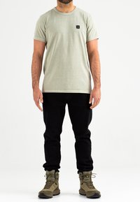 Lichtgroene T-shirt met korte mouwen en een klein zwart logo, gecombineerd met zwarte broek en groene wandelschoenen, weergegeven tegen een witte achtergrond.