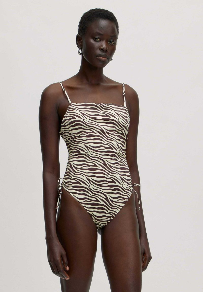 Bruin en crème zebra print badpak met verstelbare spaghettibandjes en zijkanten met strikjes. Glad materiaal met een aansluitende pasvorm.