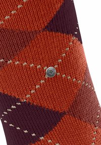 Dettaglio ravvicinato di un tessuto a maglia con motivo che presenta ricchi colori arancione, viola e crema, con un'etichetta metallica del logo visibile.