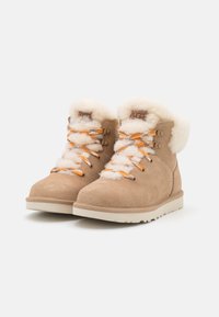 Tanfarbene Wildlederschuhe bis zum Knöchel mit weißem Futter aus Fell, orangefarbenen und grauen Schnürsenkeln und einer cremefarbenen Gummisohle; ausgestattet mit Metallaugen und geprägtem Logo.