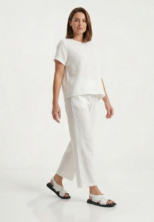 Vrouw wandelend, dragend een loszittend wit kort mouwtopje, bijpassende wijde broek en witte sandalen met zwarte zolen, tegen een effen witte achtergrond.