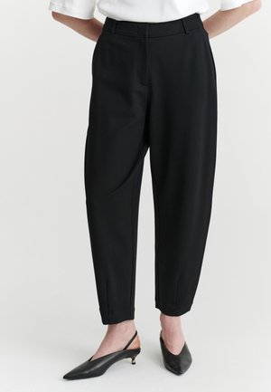 Spodná časť tela osoby, ktorá má na sebe čierne zúžené nohavice v kombinácii s čiernymi slingback lodičkami s úzkou špičkou na jednoduchom pozadí.