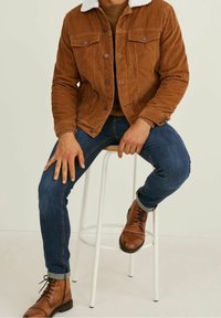 Braune Cordjacke mit einem weißen Fleece-Kragen, getragen über einem braunen Oberteil. Blaue schmale Jeans und braune Leder-Ankle-Boots. Sitzt auf einem weißen Hocker.