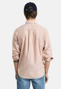  Lichtroze shirt met blauwe verticale strepen, met knoopsluiting bij de kraag, lange mouwen opgerold bij de manchetten, een plooi aan de achterkant, gedragen met blauwe jeans.