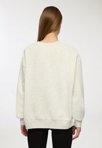 Felpa oversized grigio chiaro con collo a girocollo e polsini a costine, con una texture morbida e un orlo dritto, indossata da un modello in abbigliamento nero.